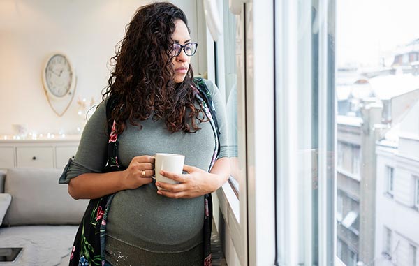 A pregnant woman standing near a window holding a cup of coffee contemplating adoption